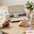 Cream Cricut Explore 4 Smart Cutting Machine crafting gold vinyl decals to personalize ceramic mugs and tags.