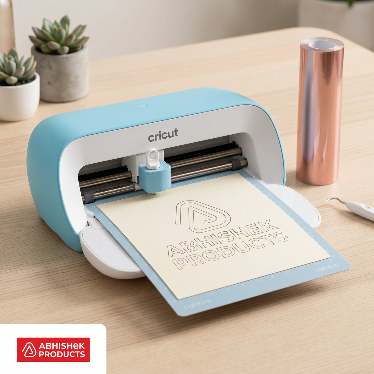 Blue Cricut Joy 2 cutting machine drawing a custom logo on cream cardstock next to a rose gold vinyl roll.